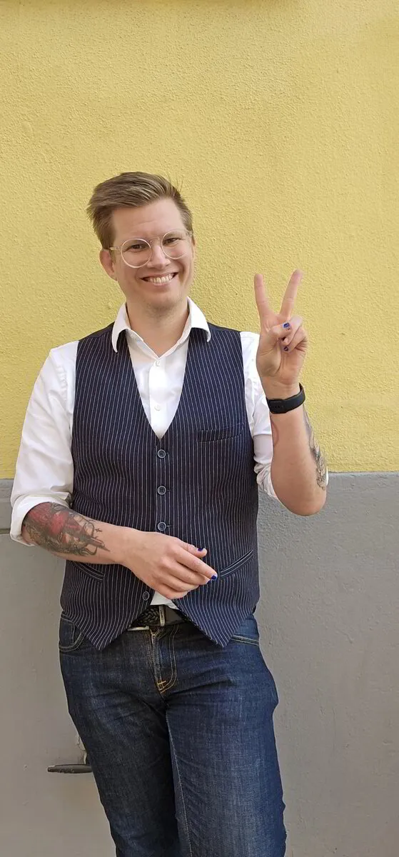 Sebastian står framför en gul vägg och gör en peace-symbol med ena handen. Han har kort hår, glasögon, en vit skjorta, mörk väst med smala ränder och jeans. Ena armen är tatuerad och han ler mot kameran.