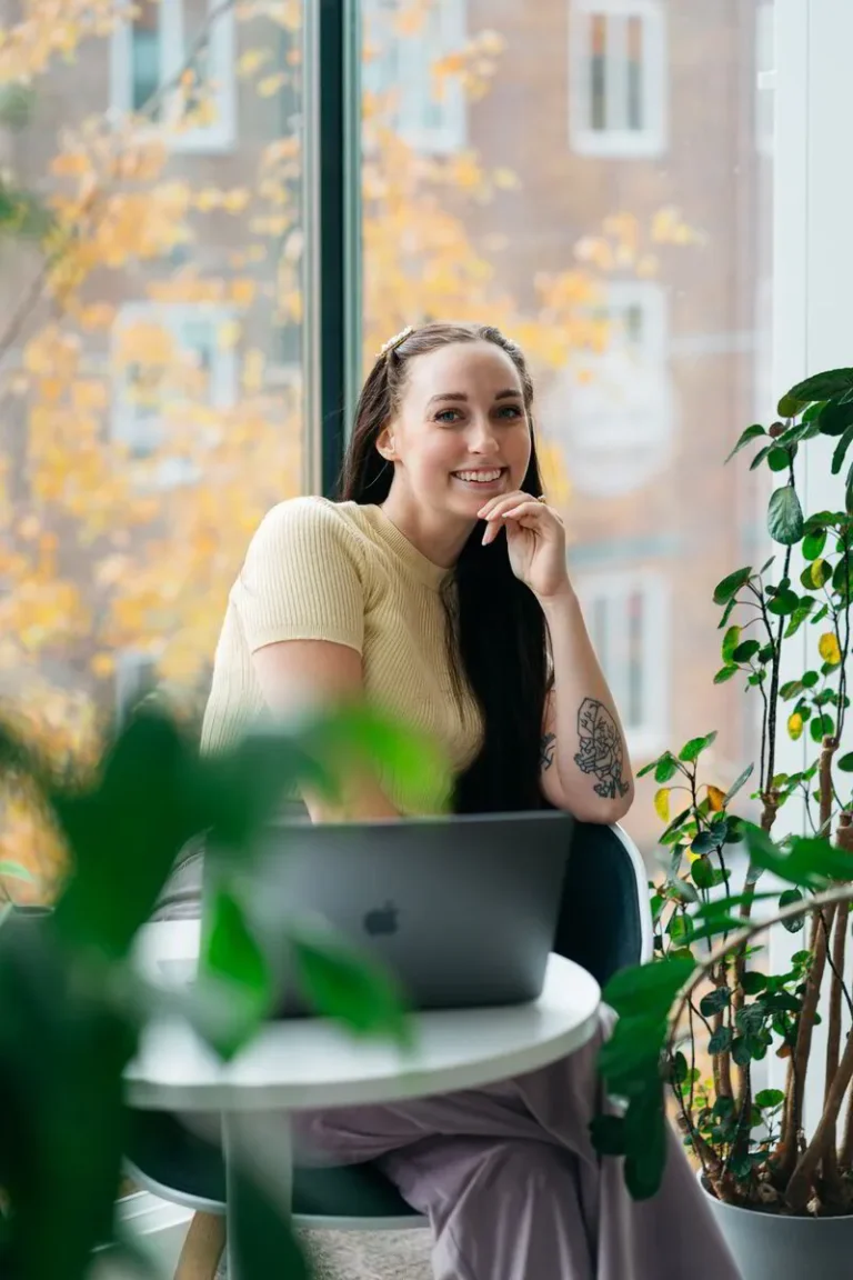 Sophie sitter vid ett runt bord med en laptop framför sig, omgiven av gröna växter. Hon har långt mörkt hår, en ljusgul stickad tröja och synliga tatueringar på ena armen. Hon ler och tittar mot kameran, med höstfärgade träd och en byggnad synlig genom det stora fönstret bakom henne.