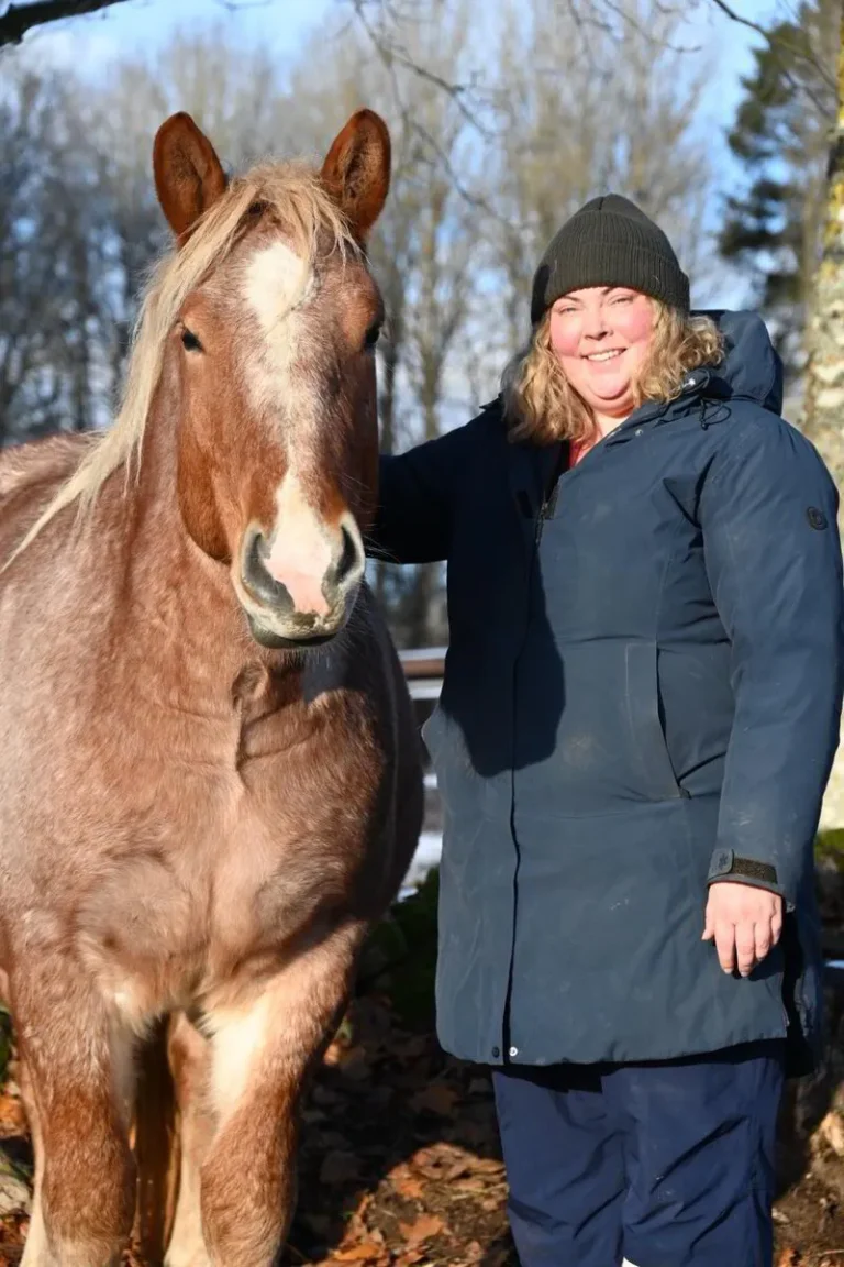 Susanna står bredvid en brun häst med ljus man en solig vinterdag. Hon ler mot kameran och har på sig en mörkblå vinterjacka och en mörk mössa. I bakgrunden syns träd utan löv och ett tunt snötäcke på marken.