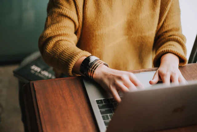 Person i senapsgul tröja arbetar på en laptop vid ett träbord. Bilden symboliserar fokus på att förbättra hemsidors design och konvertering, kopplat till Karin Bergmans webinar om hur man undviker vanliga designfällor.