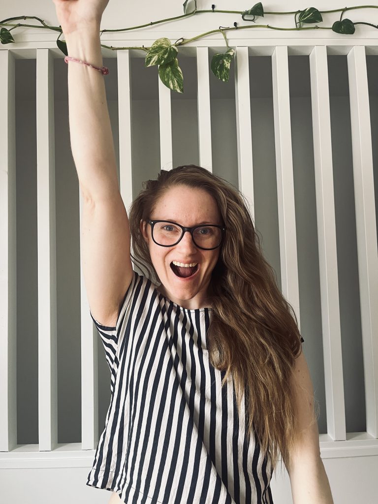 Karin Bergman ler stort och lyfter ena armen i en peppande gest framför en ljus bakgrund med vita ribbor. Bilden utstrålar glädje och energi inför lanseringen av kursen Lönsam webbdesign, som kommer att lära webbdesigners att skapa ännu mer säljande hemsidor.